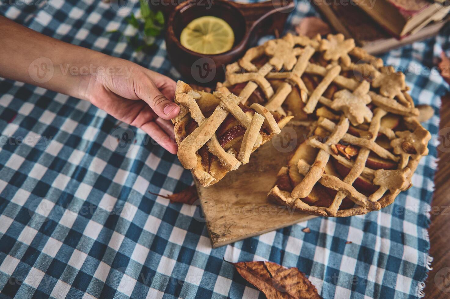Hand holding a slice of homemade apple pie on a wooden board. The pie is on a blue and white checkered tablecloth, surrounded by autumn leaves and a lemon slice in a wooden bowl. photo