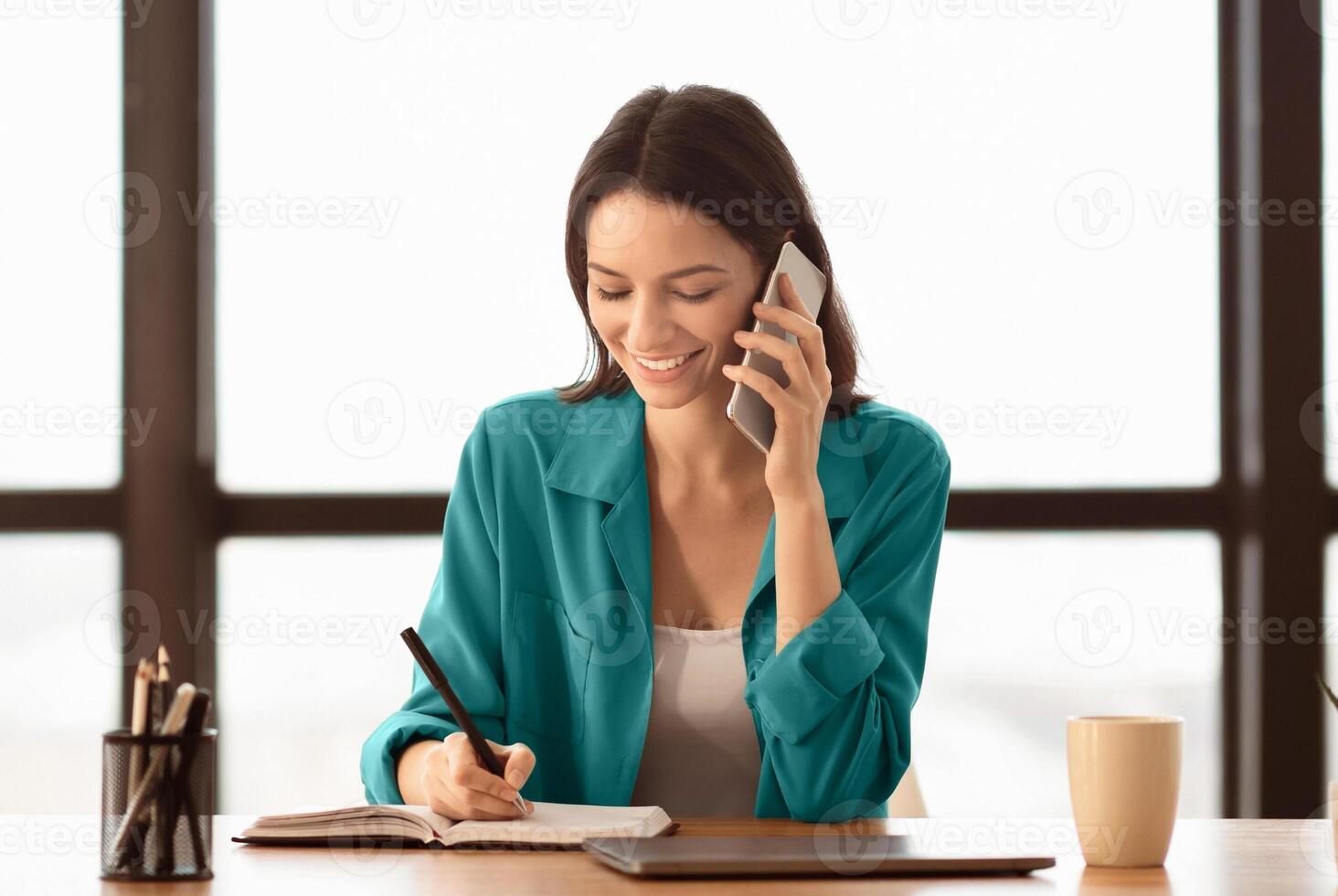 Smiling business woman talking on phone and taking notes, sitting at her working place in office, copyspace photo
