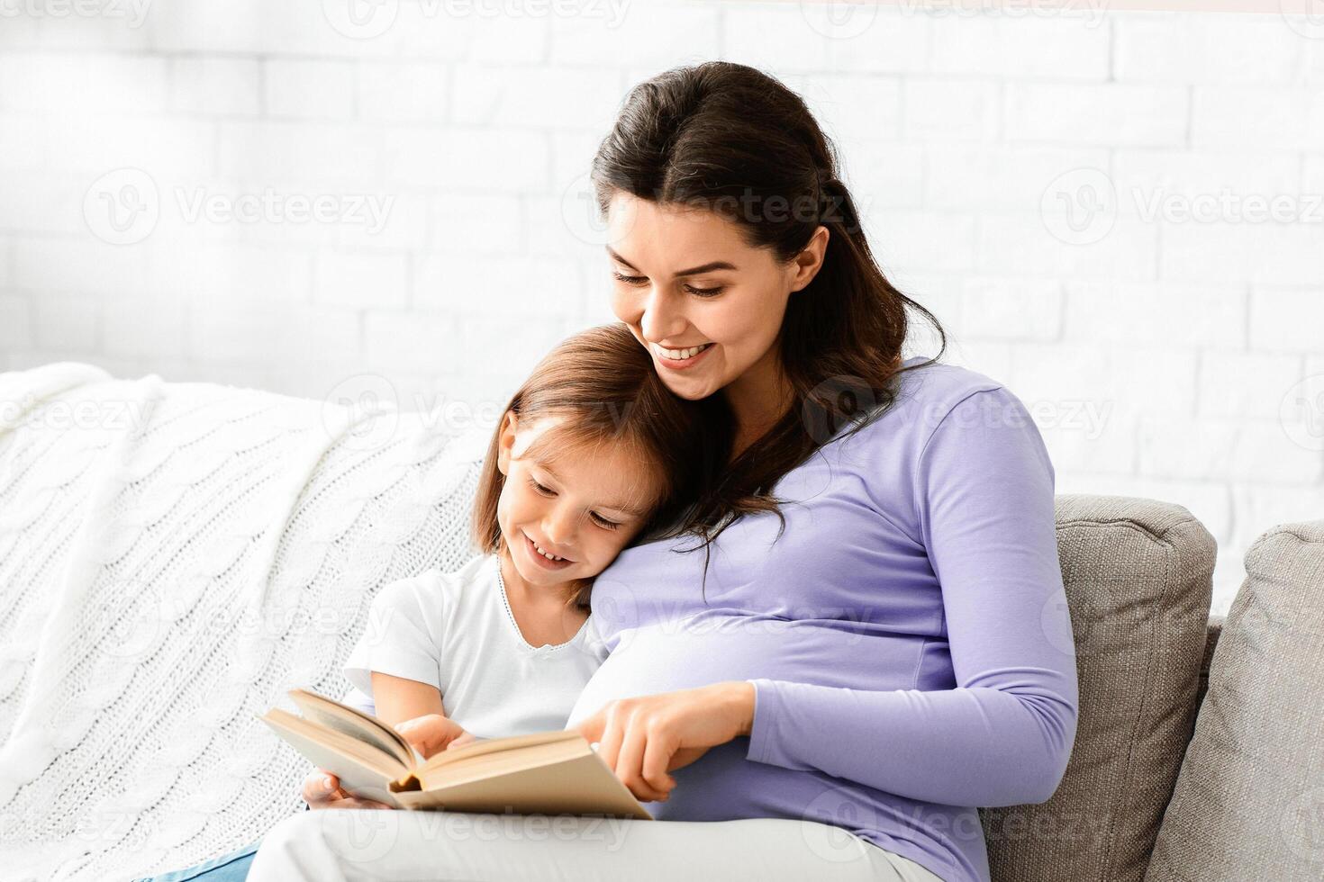Friendly intelligent family pregnant mother and little daughter reading book on couch at home, empty space photo
