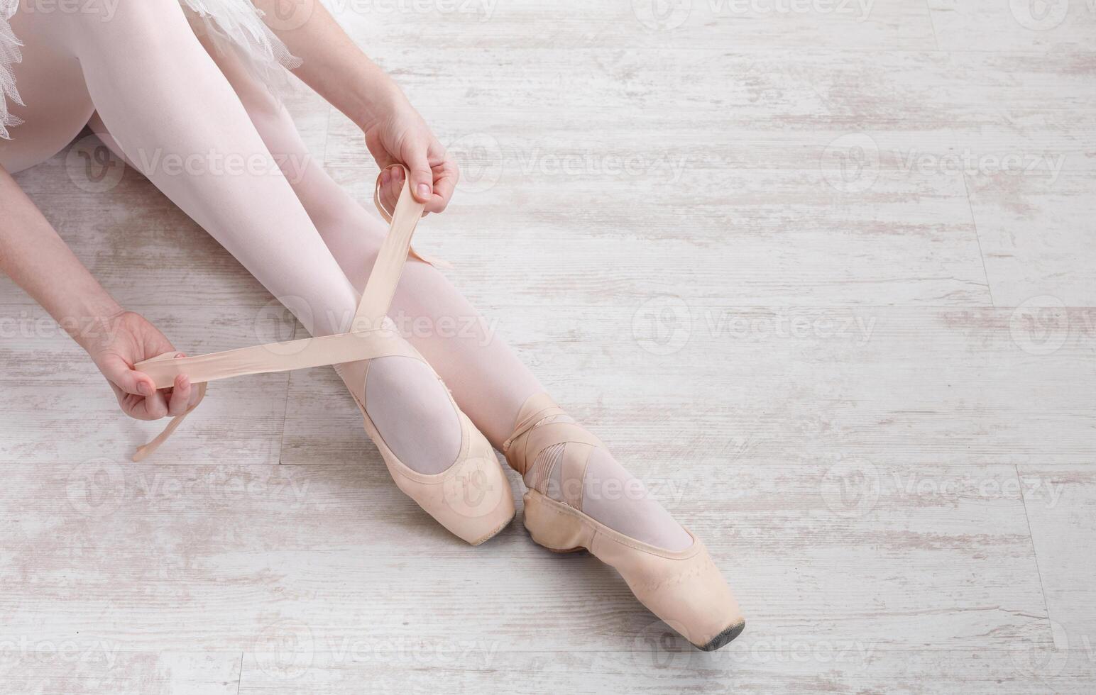 Beautiful legs of young ballerina who puts on pointe shoes at white wooden floor background, top view from above with copy space. Ballet practice. Beautiful slim graceful feet of ballet dancer. photo