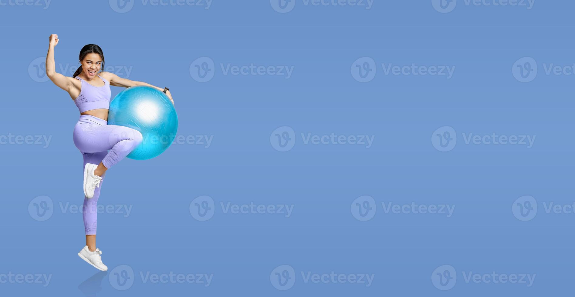 A young woman energetically performs an exercise while balancing on a fitness ball, showcasing her enthusiasm in a bright blue studio. She wears athletic clothing and exudes confidence. photo