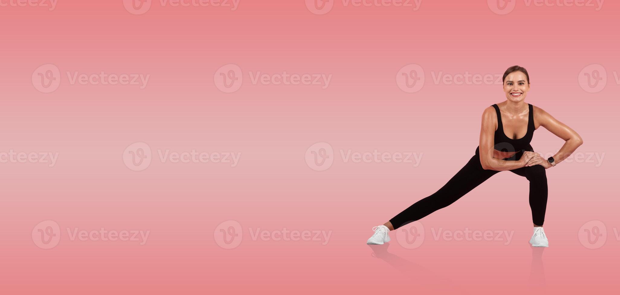 A woman in athletic wear performs a side lunge, showcasing flexibility and strength. The vibrant pink background adds energy to this workout setting. photo