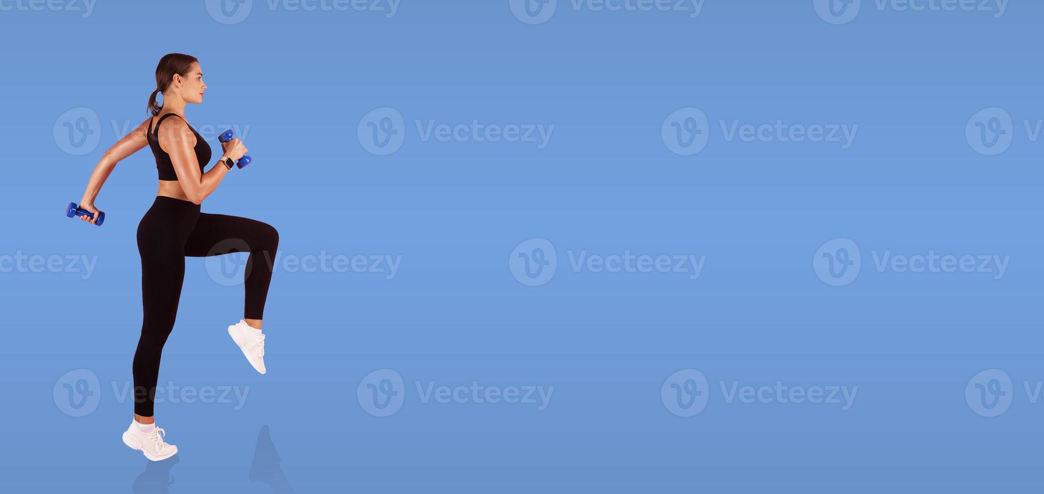 A woman engages in a fitness routine, lifting dumbbells while performing a leg exercise. She wears athletic clothing and focuses on her movement against a solid blue background. photo