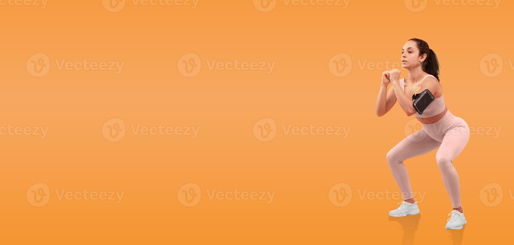 A woman in workout attire is engaged in a squat exercise, showcasing her form in a bright orange studio. The energetic environment emphasizes her dedication to fitness. photo
