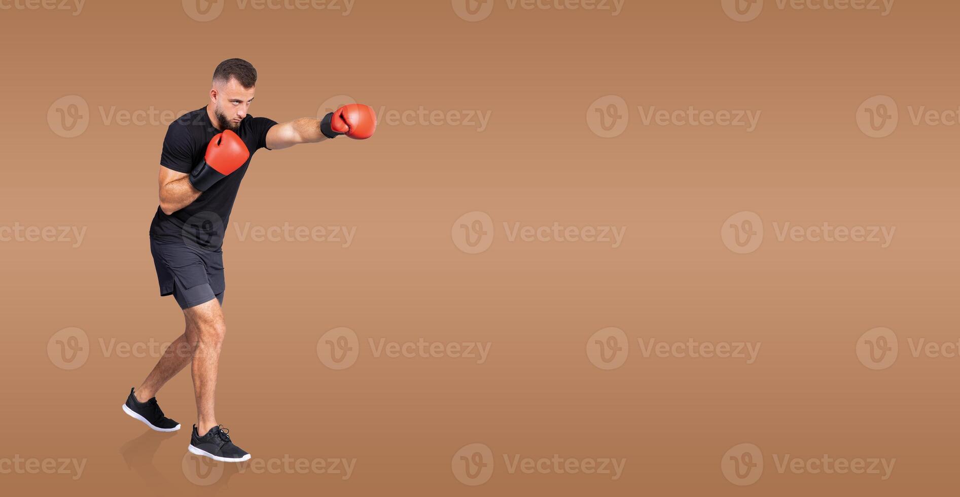 A focused man boxer in training throws a punch while wearing red boxing gloves. He is dressed in casual athletic wear, showcasing his dedication and skill in a well-lit indoor setting. photo