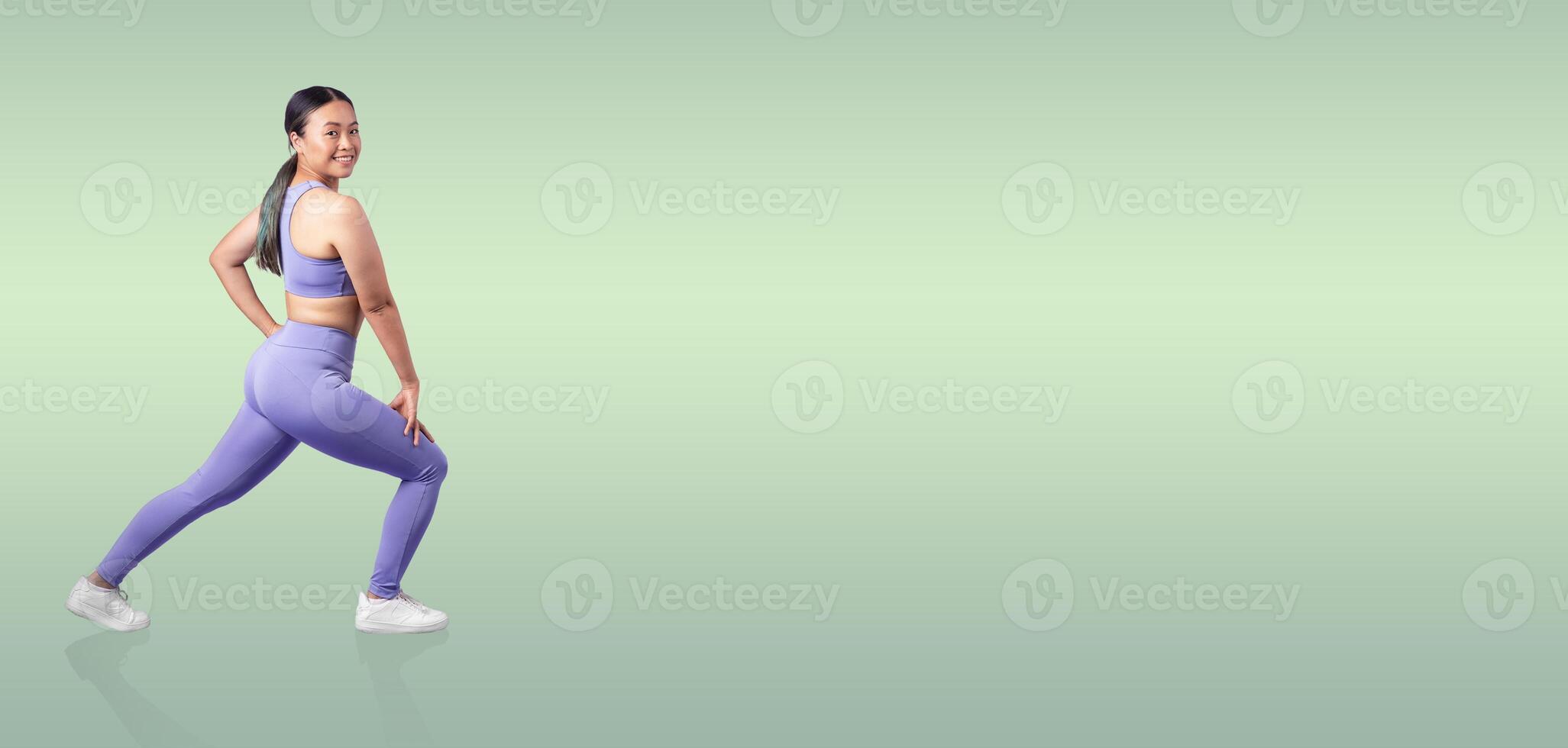 A fit woman performs a lunging exercise while wearing stylish activewear. She smiles confidently against a soft, soothing green background, promoting fitness and health. photo