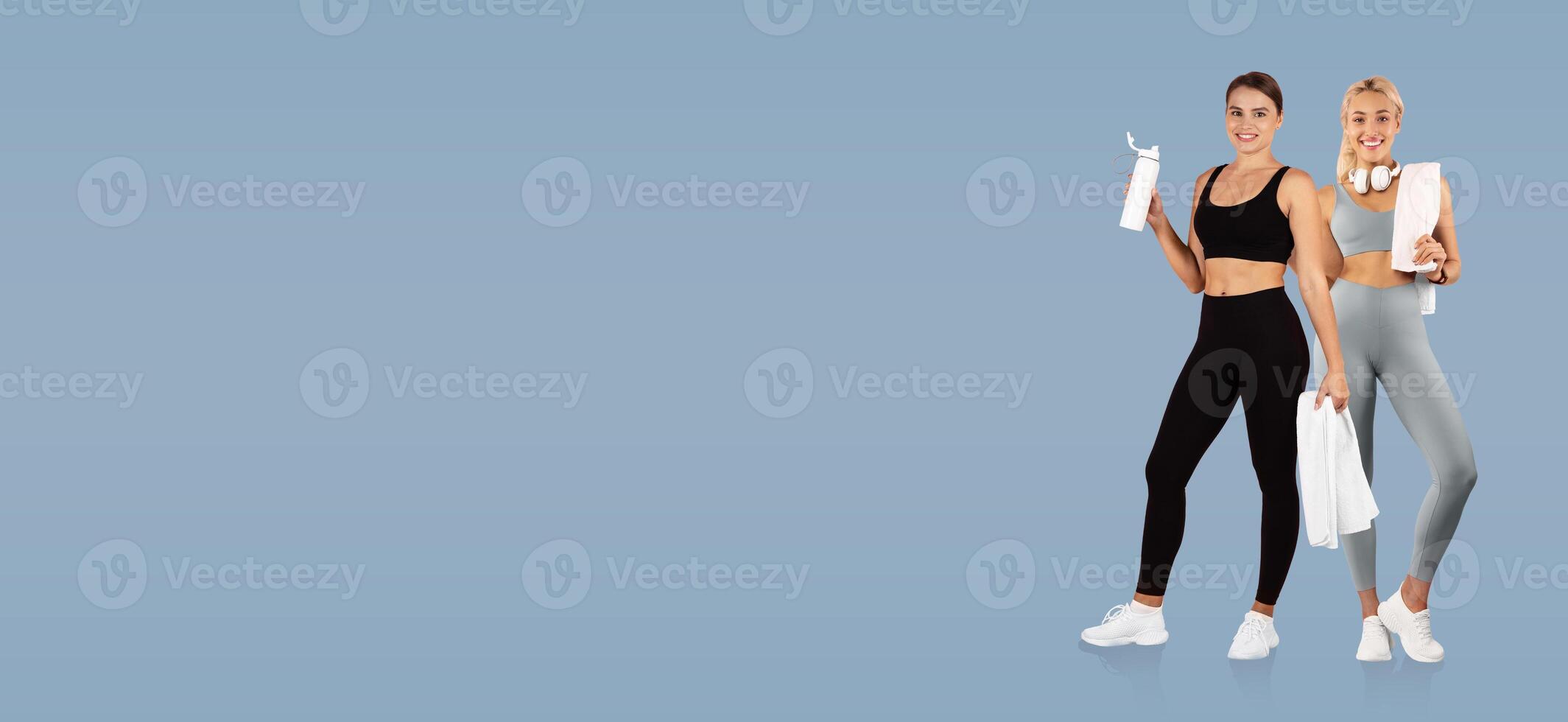 Two women in athletic wear stand against a blue background, each holding water bottles and towels while showcasing their fitness-focused attitudes and style. photo