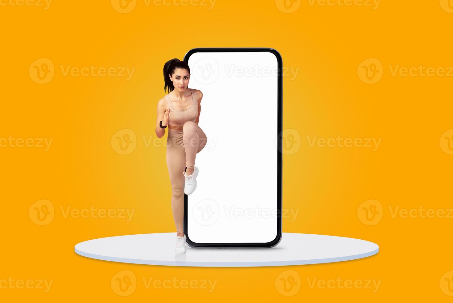 A woman in minimal activewear performs a dynamic exercise nearby a large smartphone model against a bright yellow background. The energetic pose suggests a fitness-focused theme. photo