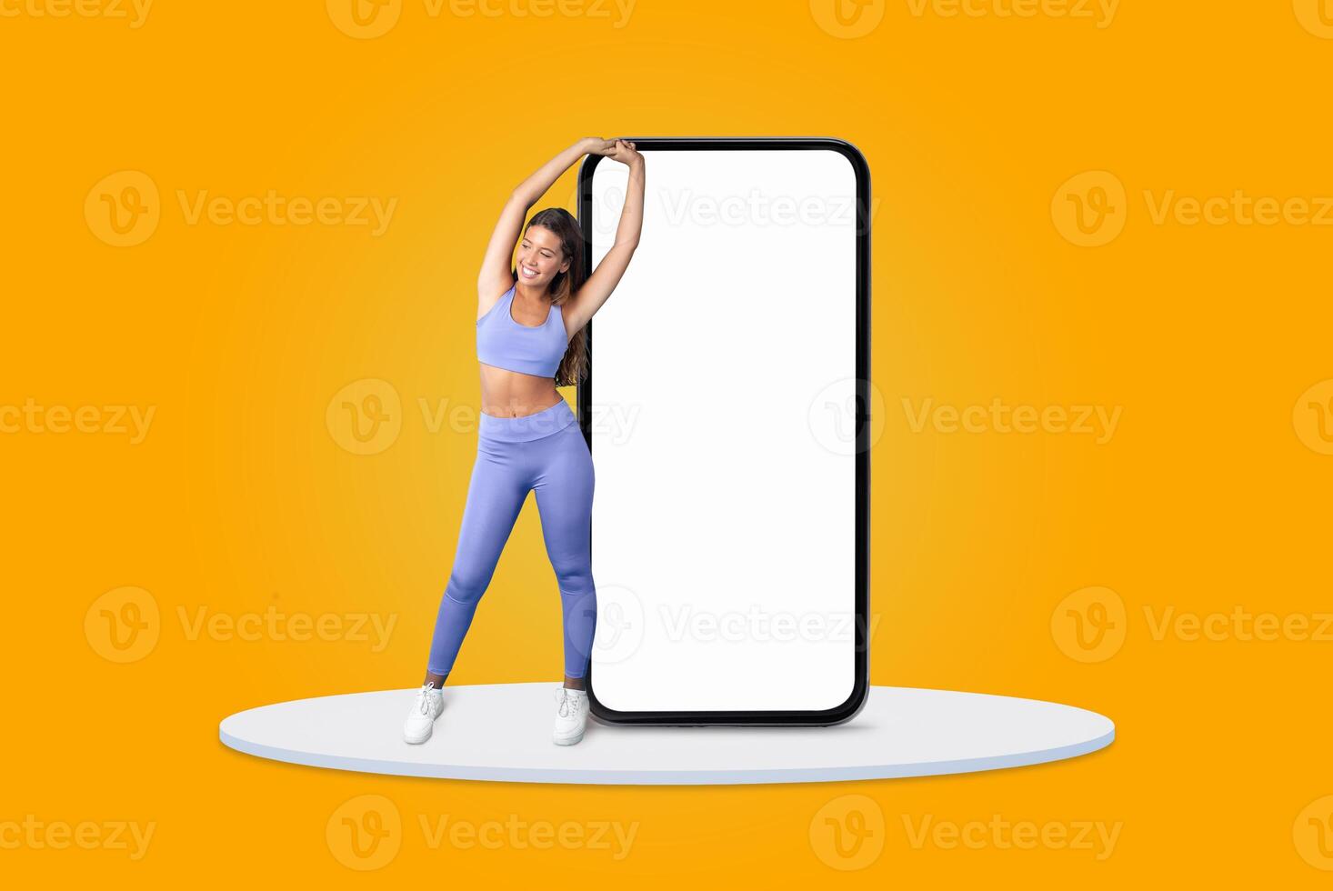 A young woman dressed in purple activewear stands beside an oversized blank smartphone. She smiles confidently while posing against a bright yellow background, showcasing fitness and enthusiasm. photo