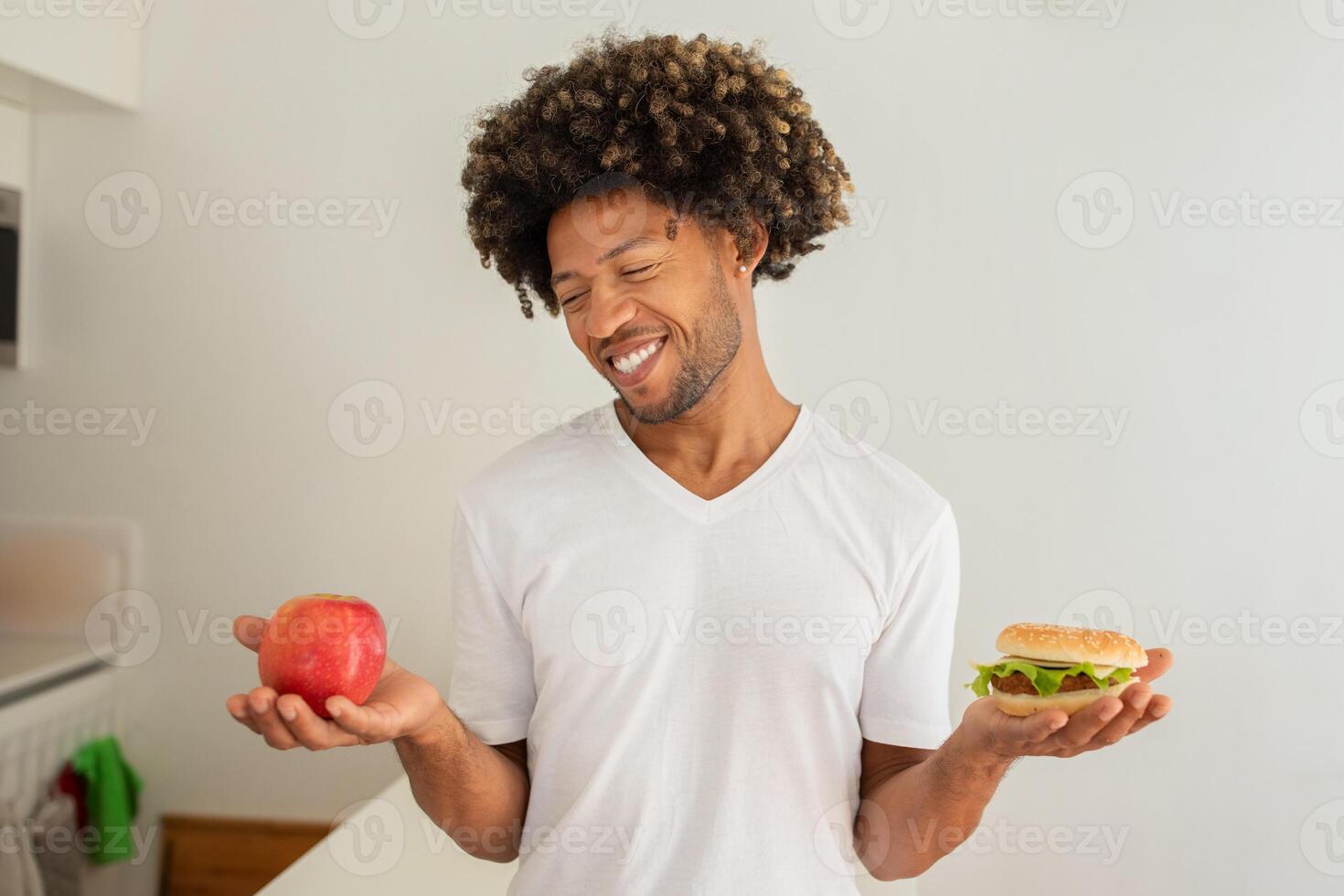 A black man stands in a well-lit kitchen, holding an apple in one hand and a burger in the other, contemplating his food choices with a smile on his face. photo
