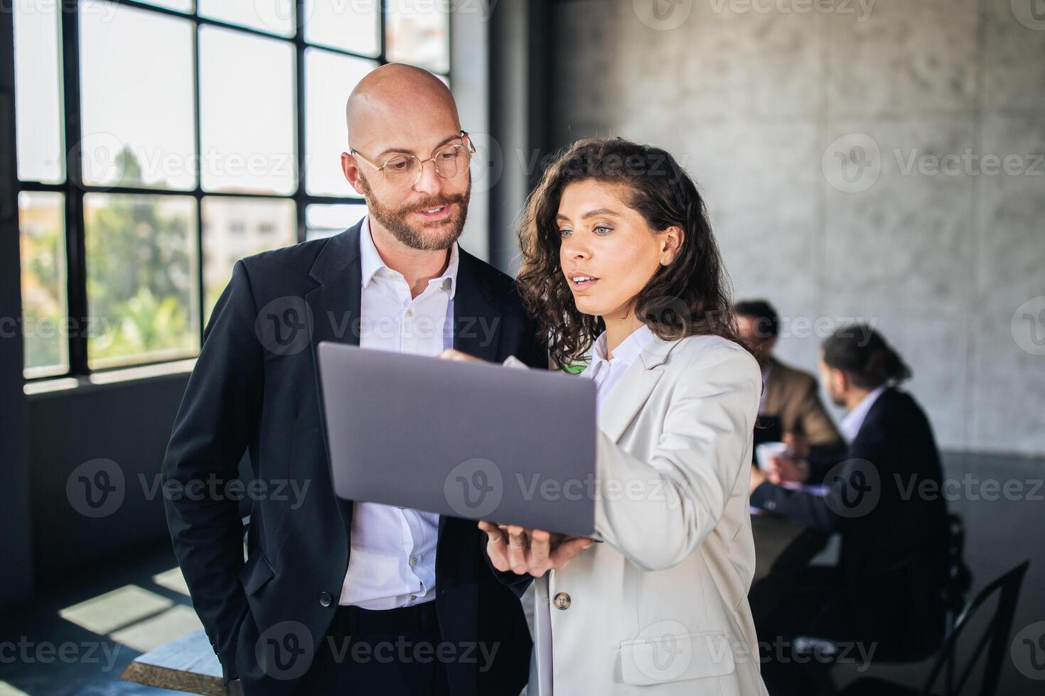 Two colleagues engage in a collaborative discussion while reviewing a laptop. The backdrop features a contemporary office with a large window and a team in the background. photo