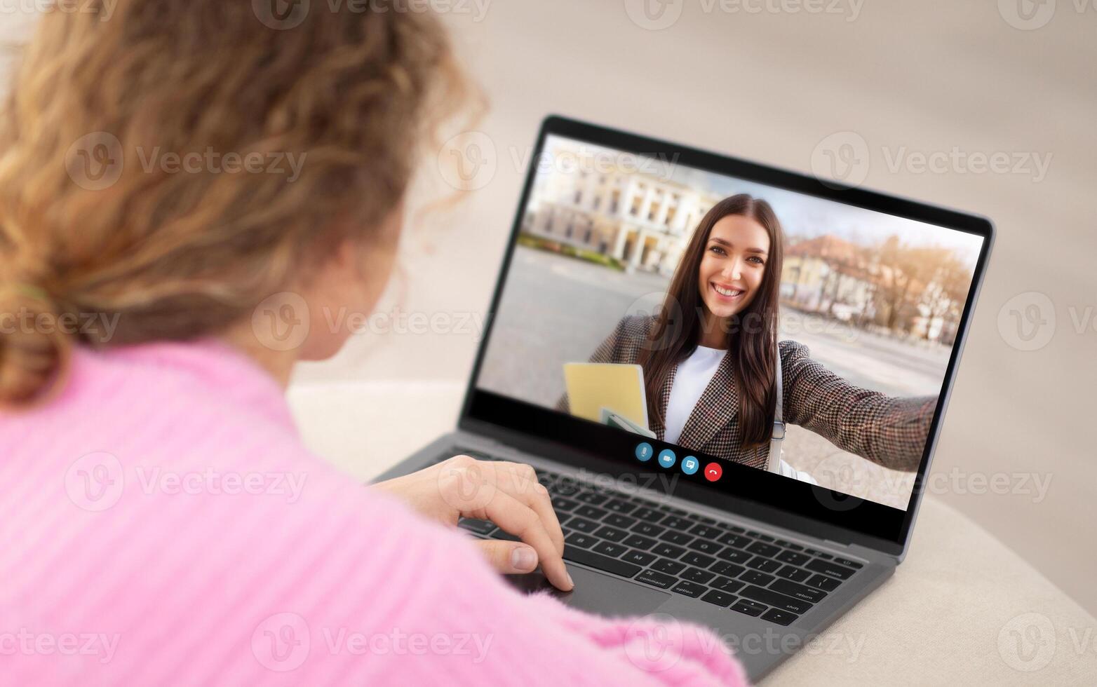 A woman interacts with a friend on a call, smiling and holding a yellow envelope. She sits at a table, with a laptop displaying her friend's cheerful face and a blurred outdoor background. photo