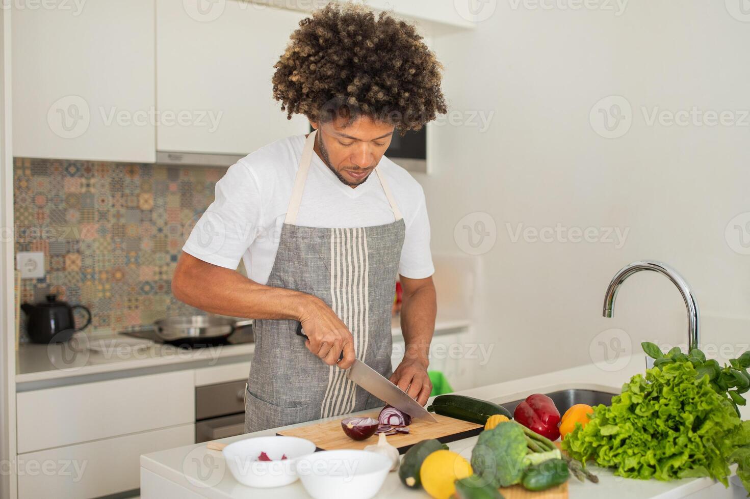 A black man is skillfully chopping vegetables on a wooden cutting board in a well-lit kitchen, surrounded by fresh produce. He is focused on preparing a healthy meal. photo
