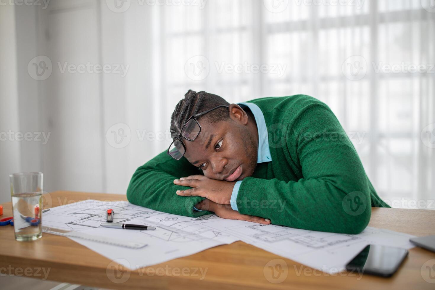 A young black man, wearing glasses looks tired and upset as he rests his head on a desk littered with blueprints and stationery in a well-lit workspace during the day. photo