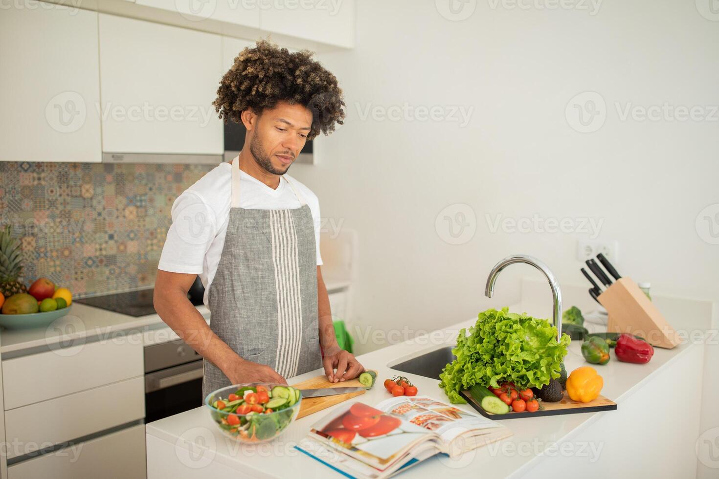 A black man with curly hair is chopping vegetables while standing in a bright, modern kitchen. He is focused on creating a fresh salad, surrounded by colorful ingredients. photo