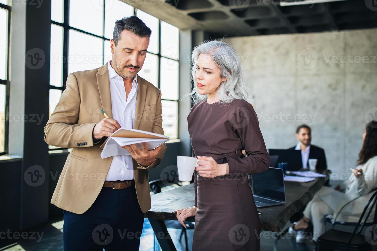 Two colleagues engage in a focused discussion while reviewing notes in a contemporary office. Others work in the background, contributing to a collaborative atmosphere. photo
