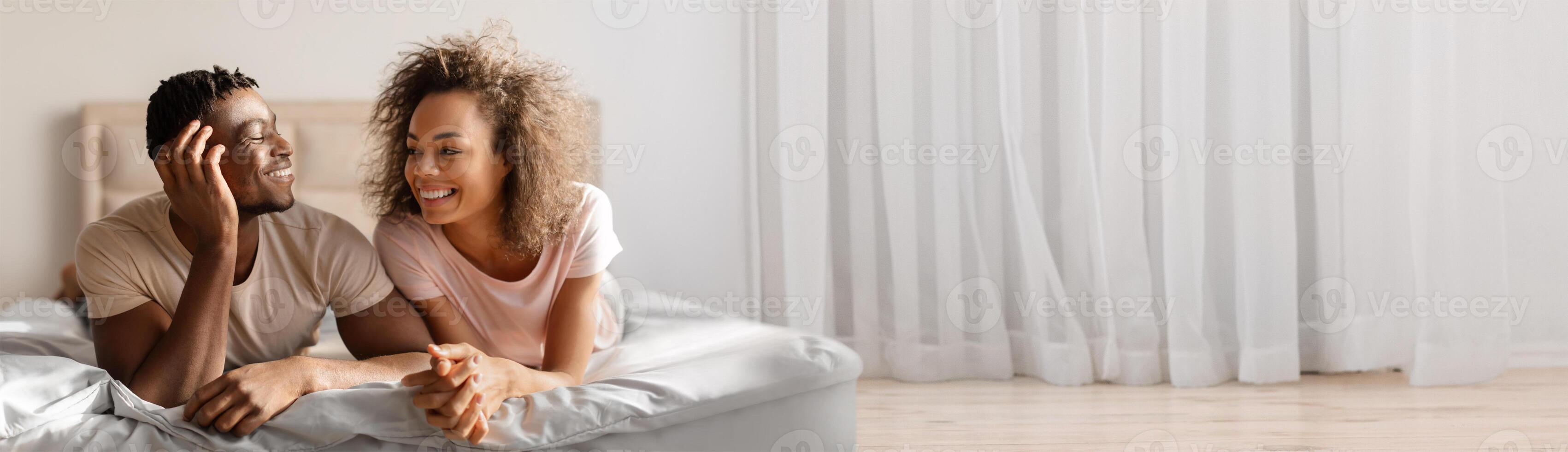 Black couple relaxes and smiles at each other on their bed, creating a warm, intimate atmosphere in the well-lit bedroom. Sunlight filters through sheer curtains, enhancing their connection. photo