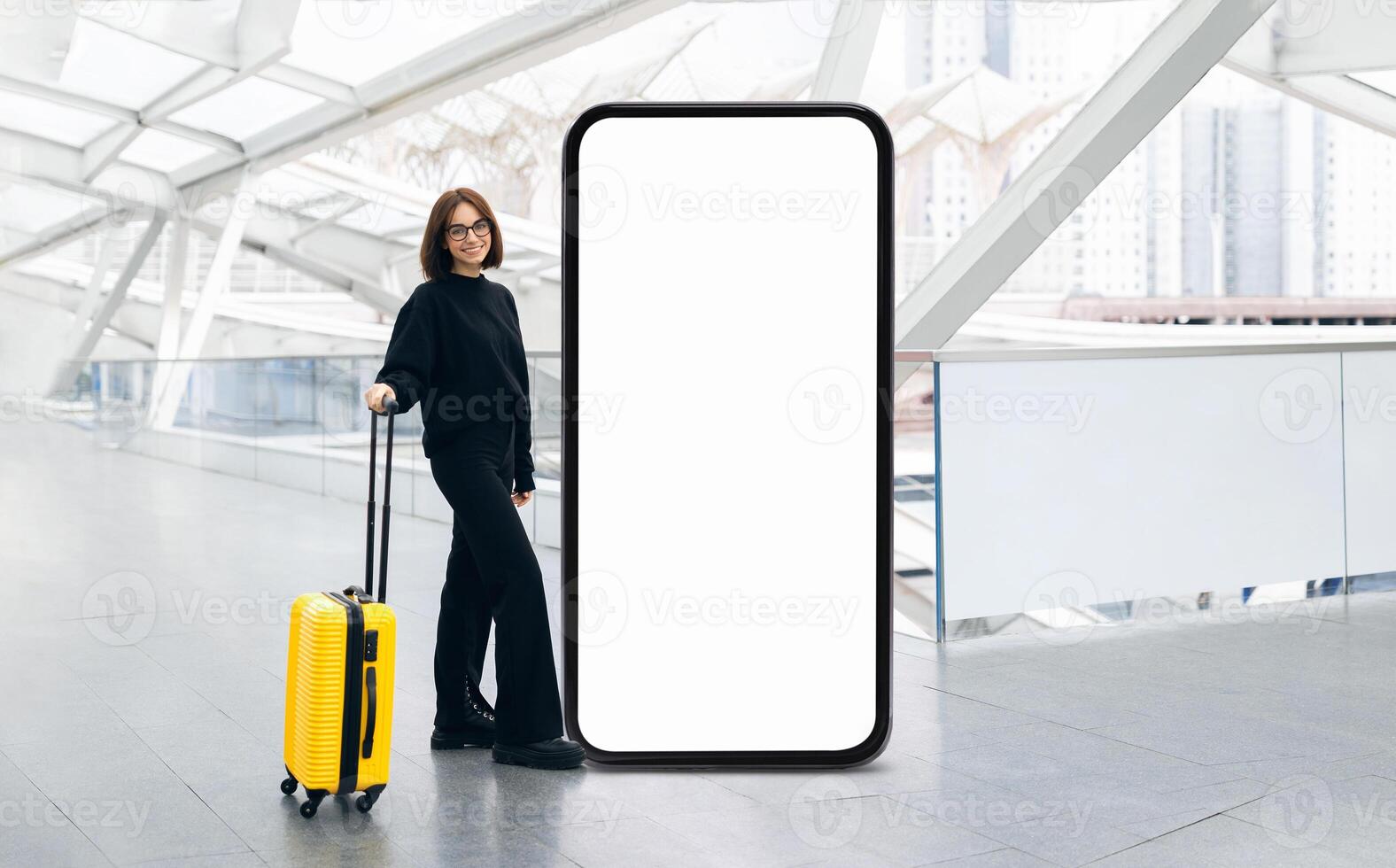 A traveler stands proudly with a yellow suitcase next to an oversized blank smartphone display in a contemporary airport terminal photo