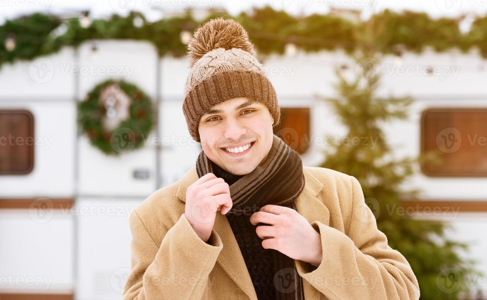 A cheerful young man wearing a knitted hat and scarf stands outside a festive mobile home, surrounded by snow, displaying a warm smile against a winter backdrop. photo