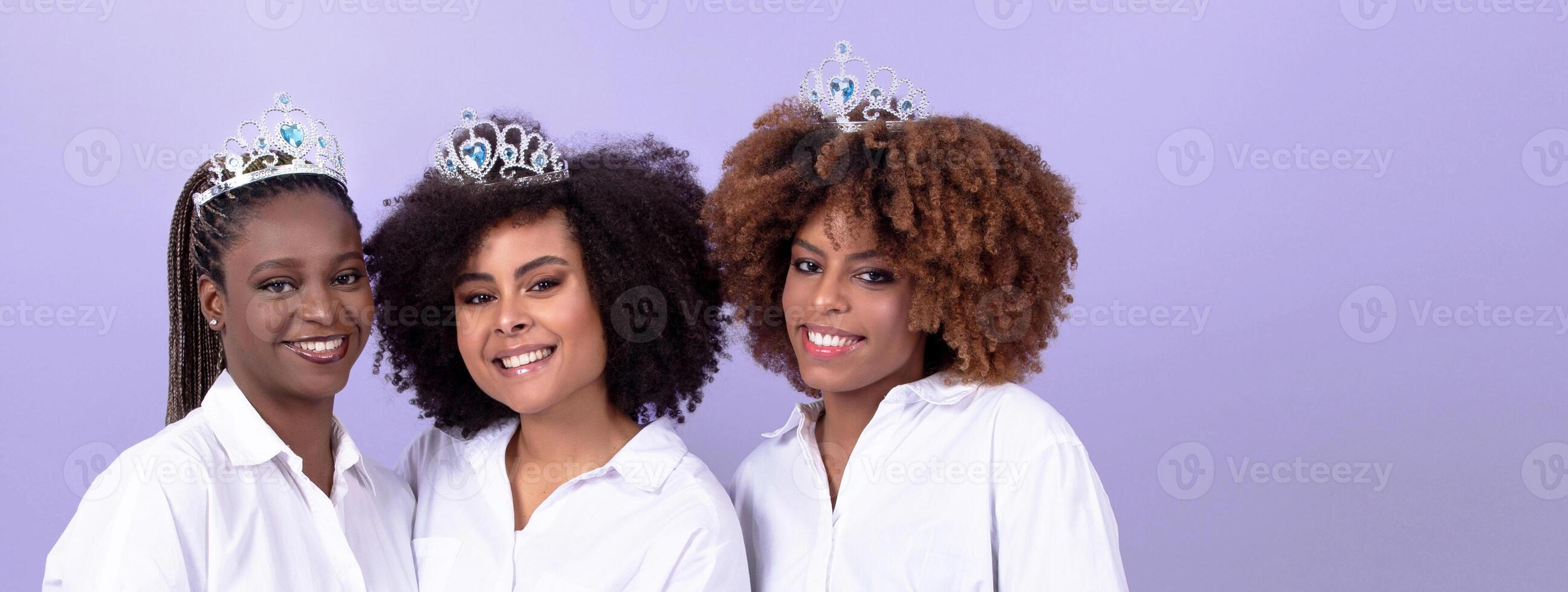 Three black women with beautiful, diverse hairstyles and crowns smile joyfully towards the camera, showcasing their unique looks and vibrant energy in a playful setting photo