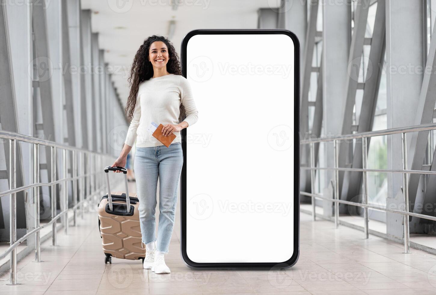 A young woman is walking confidently in a modern corridor, pulling her suitcase behind her while holding a travel document. A large blank screen stands beside her, creating a striking backdrop. photo