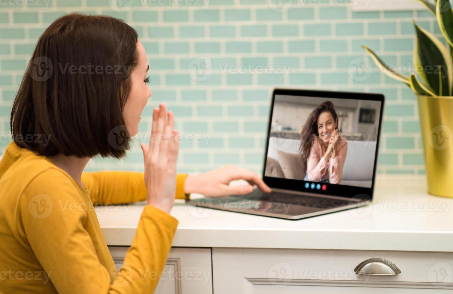 A woman wearing a yellow top smiles and waves at her friend during a call, sitting at her kitchen table with modern decor and a plant nearby. photo