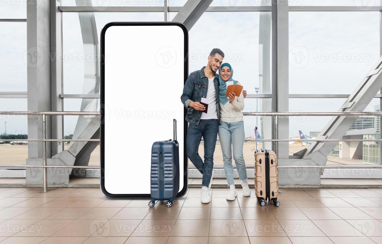 A cheerful muslim couple stands together by huge phone with blank screen at the airport, with luggage nearby, while they engage with their devices photo