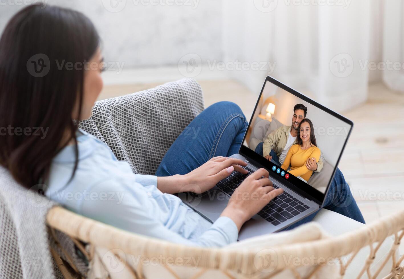 A woman engaged in a call on her laptop, smiling at a couple on the screen. She is seated in a cozy chair, with a bright, airy room in the background. photo