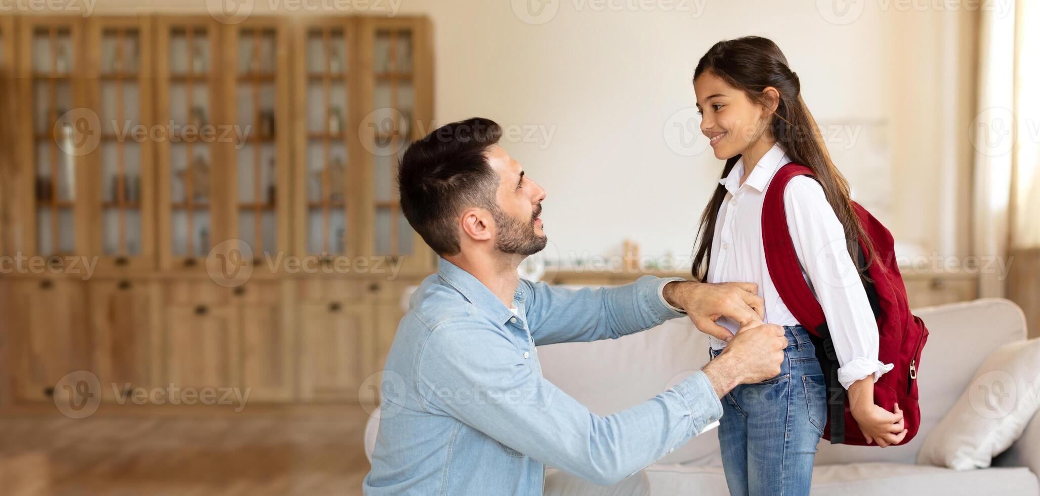 A father is adjusting his daughter backpack as she stands smiling. The warm and inviting atmosphere of their living room highlights the bond they share before school. photo