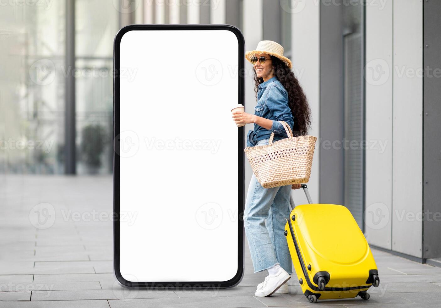 A young woman stands in a sleek urban plaza, featuring a large blank screen. She carries a yellow suitcase and a woven basket, enjoying her coffee while embracing her travel adventure. photo