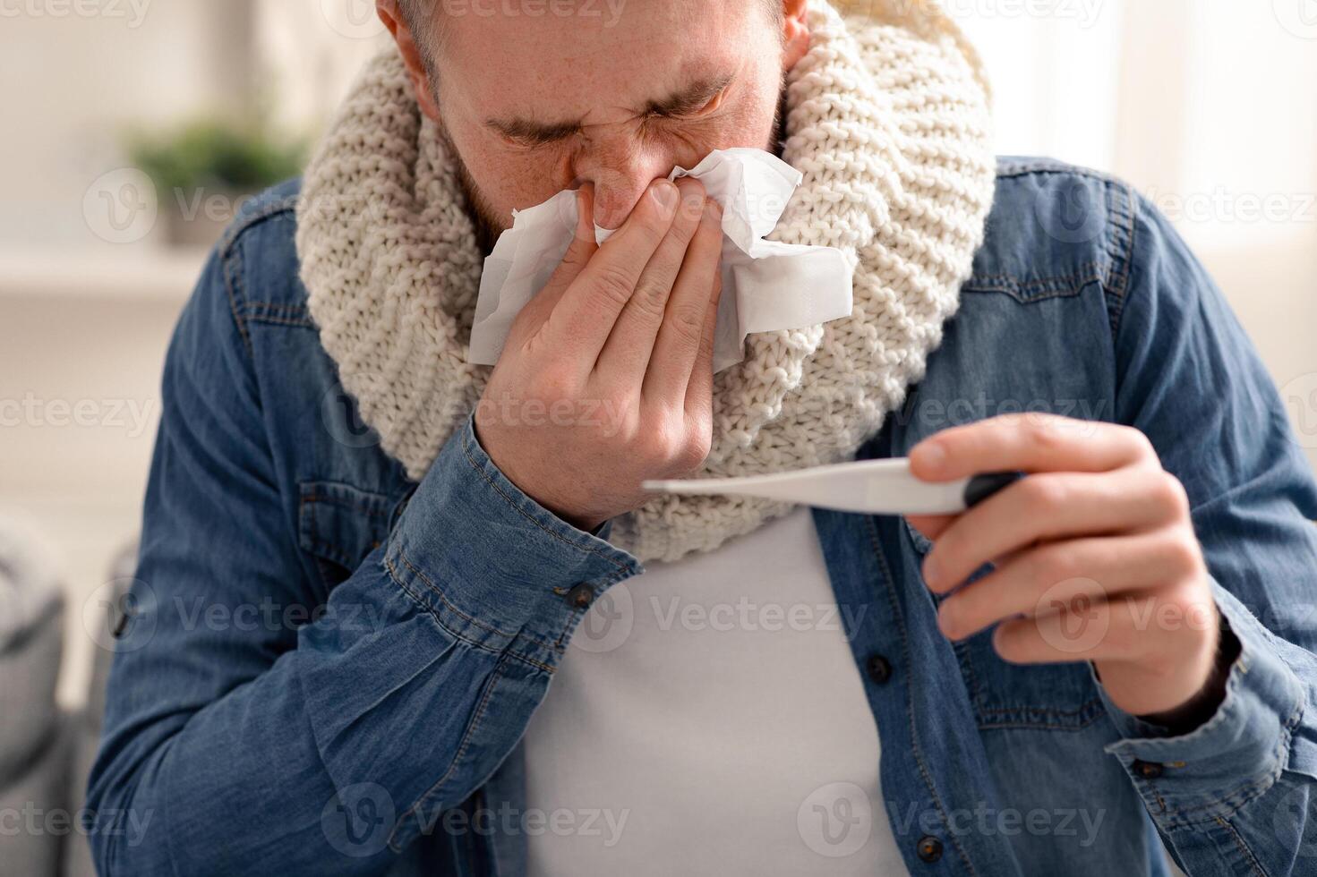 Seasonal flu. Sick man checking his body temperature and blowing nose at home photo