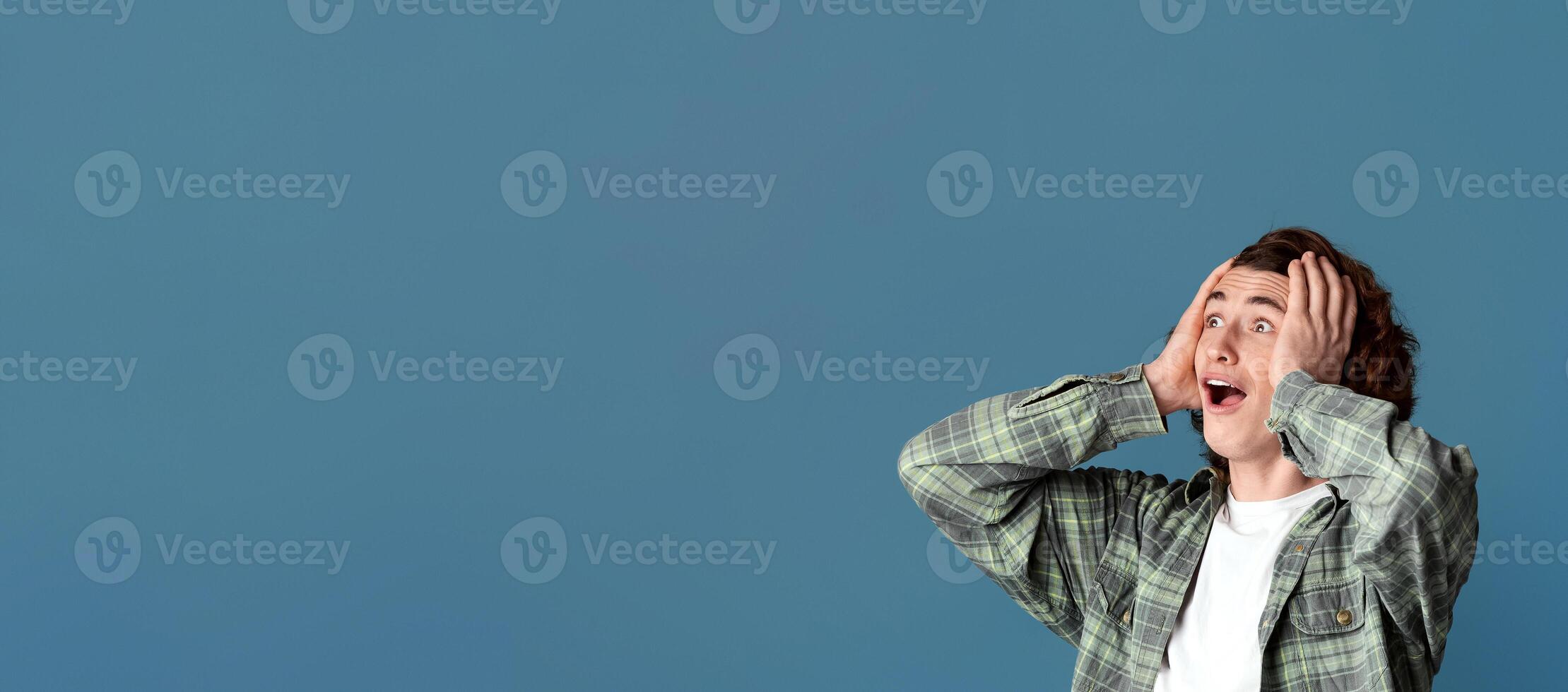 Excited millennial guy holding his head and shouting OMG on turquoise studio background, copy space photo