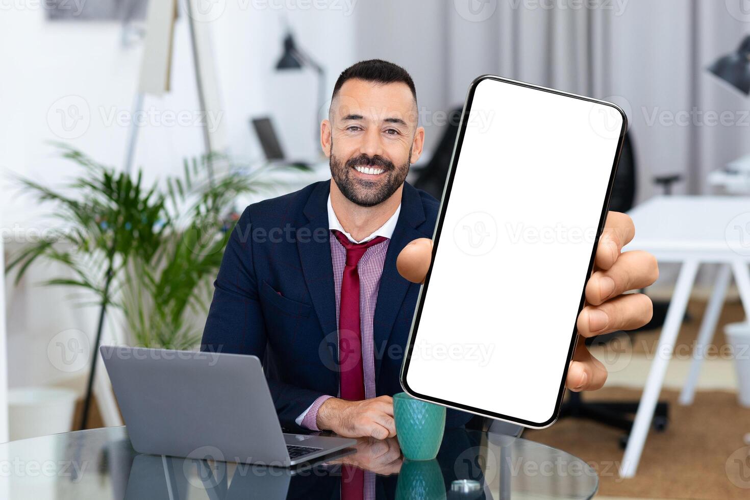 A man in formal attire sits at an office desk, smiling and holding a smartphone in front of him. His laptop and a cup are nearby, creating a productive atmosphere. photo