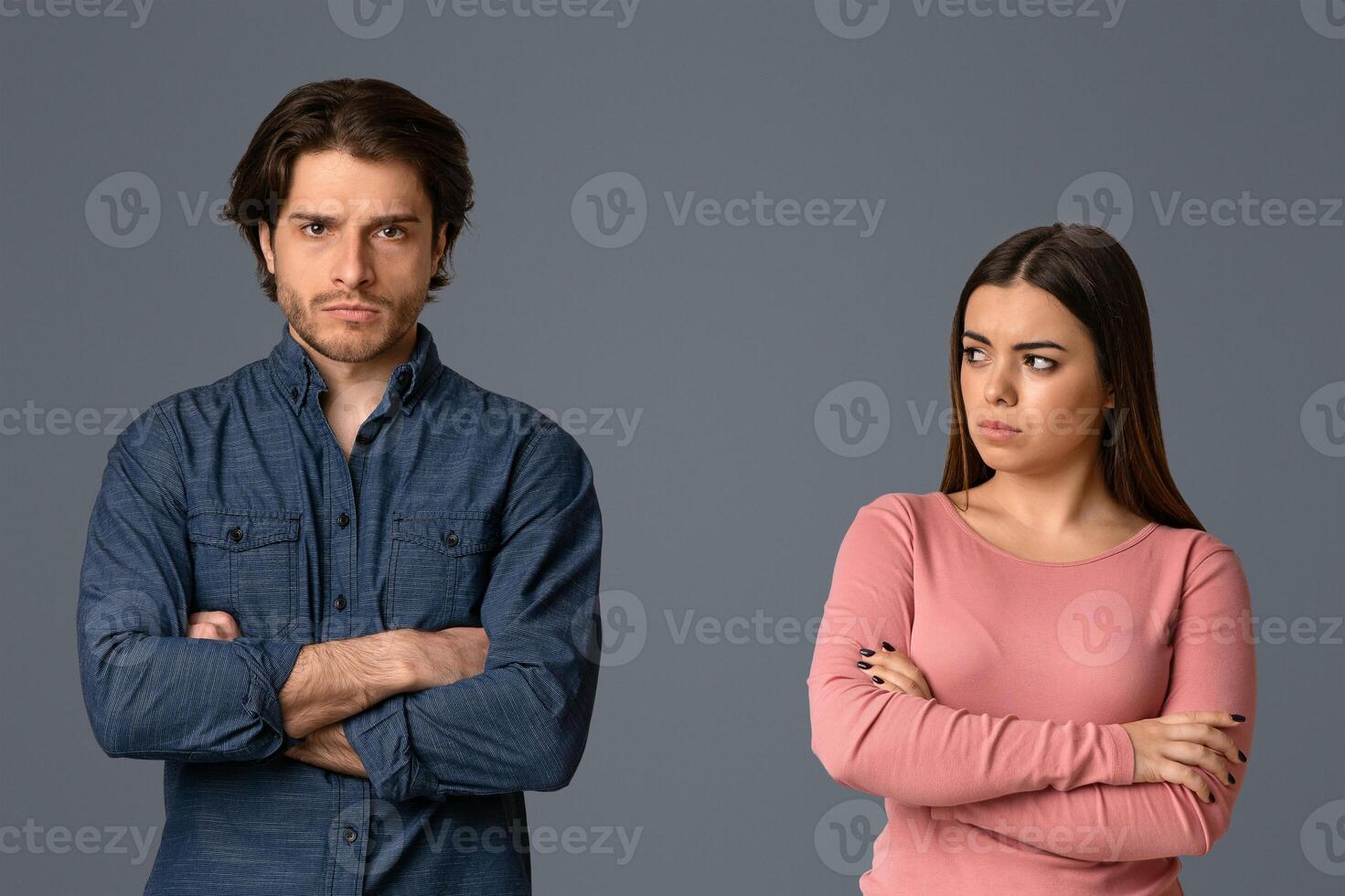Young woman looking with sorrow at her offended husband after their argue, couple standing with folded arms over grey background, empty space photo