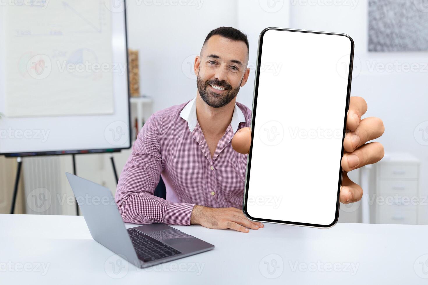 A man with a friendly smile holds up a blank smartphone in a bright office. A laptop is placed beside him, creating a workspace ambiance suitable for digital activities. photo
