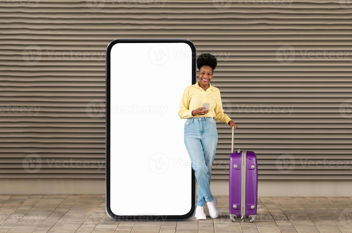 A cheerful young black woman stands next to a large blank smartphone display, holding a mobile device. She has a purple suitcase and is dressed casually, enjoying the outdoor atmosphere. photo