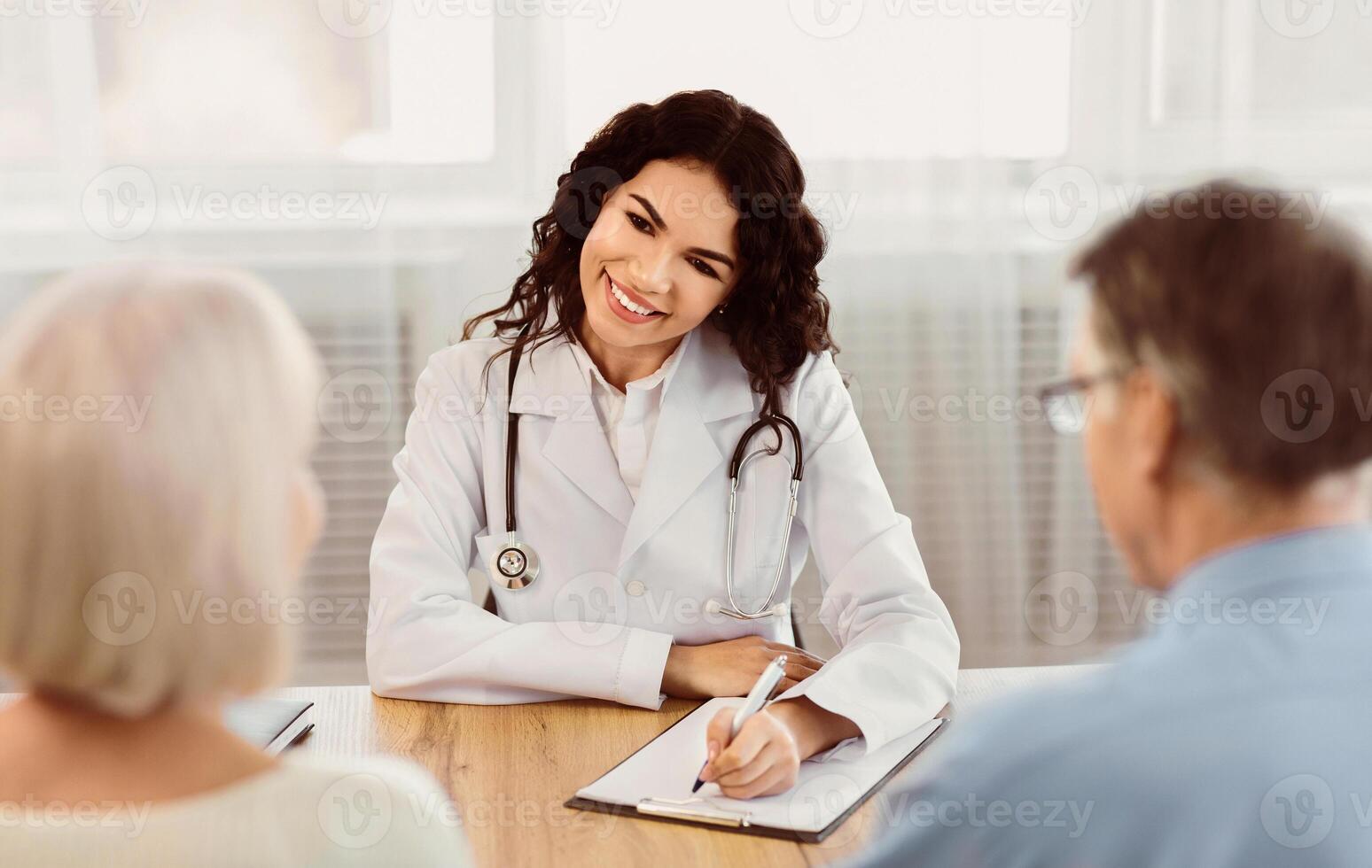 Healthcare Concept. Happy latina doctor taking notes about symptoms, filling out medical prescription to mature couple photo
