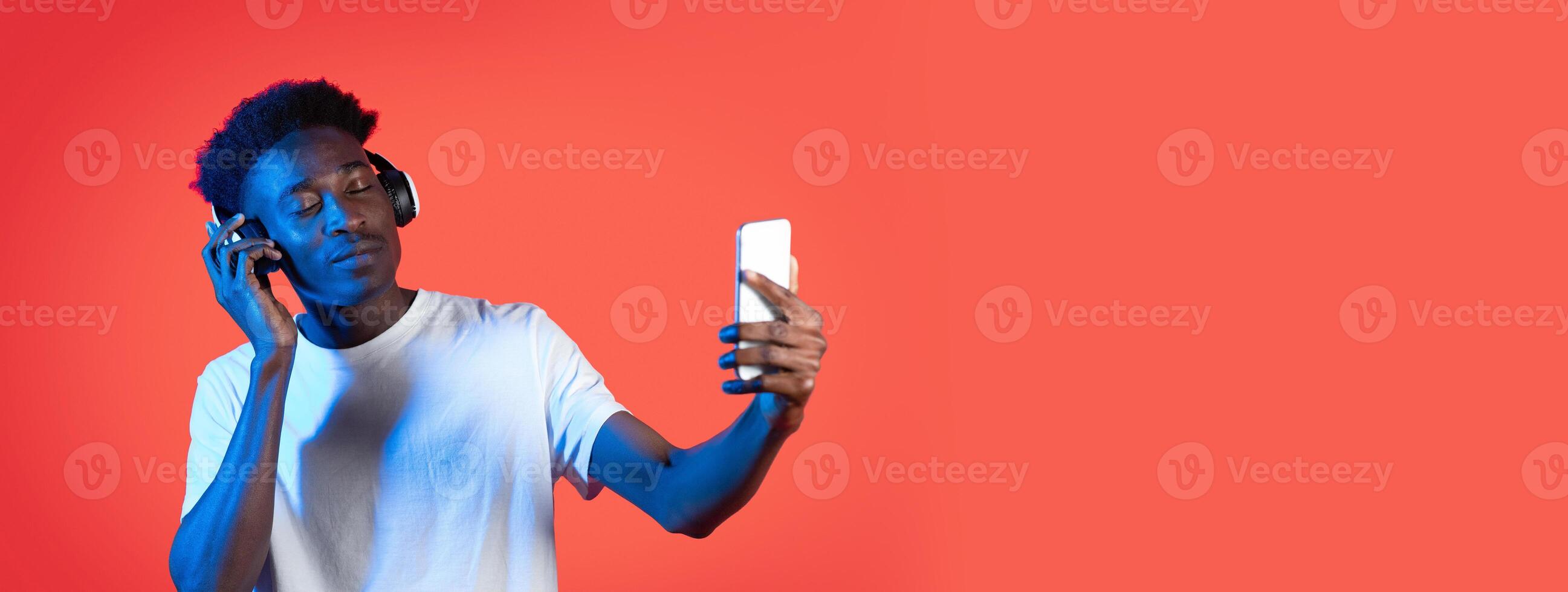 A young man wearing headphones enjoys music while taking a selfie with his smartphone. The bright solid-colored background adds vibrancy to the moment, capturing his relaxed expression. photo