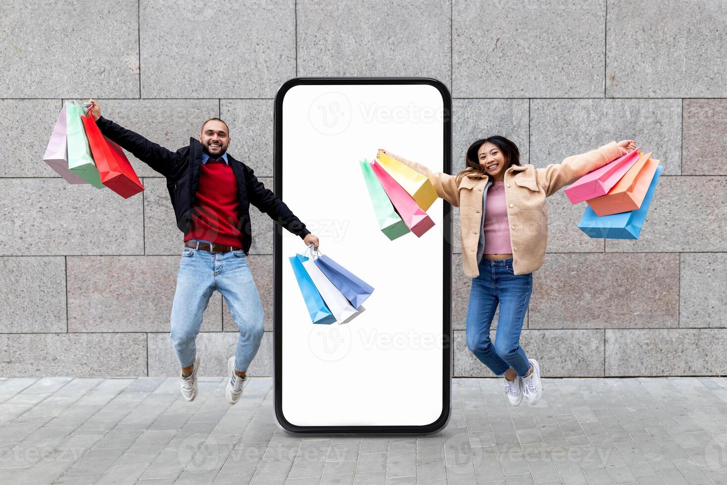 A young couple jumps in excitement while holding colorful shopping bags in front of a large blank smartphone. They convey joy and enthusiasm in their retail experience. photo