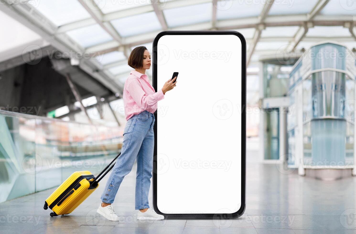 A traveler stands in an airport terminal, checking their phone while leaning against a large blank advertisement display. A yellow suitcase rests beside them, adding color to the scene. photo