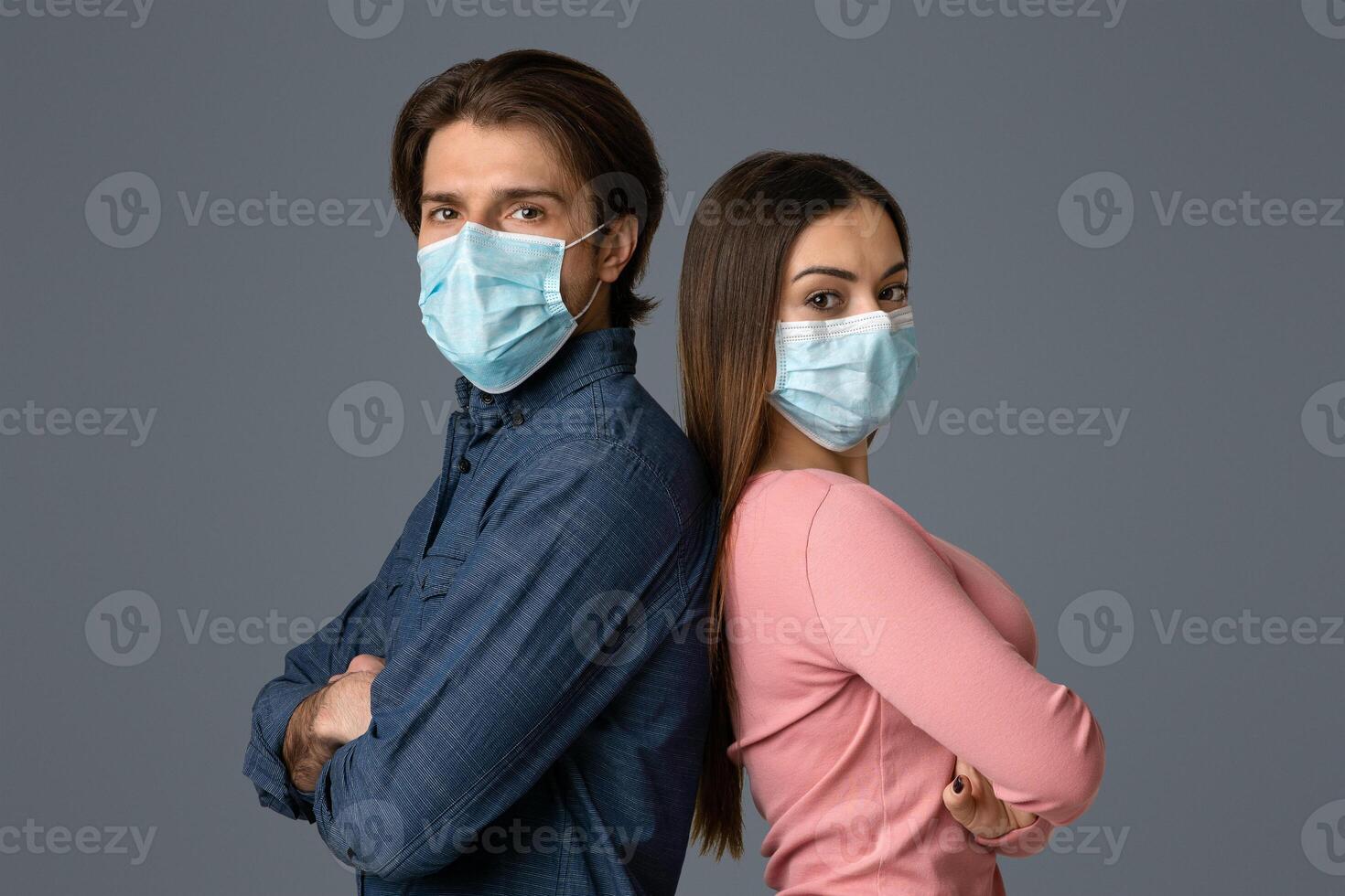 Confident Young Man And Woman In Protective Medical Masks Standing Back To Back With Folded Arms Over Light Background photo