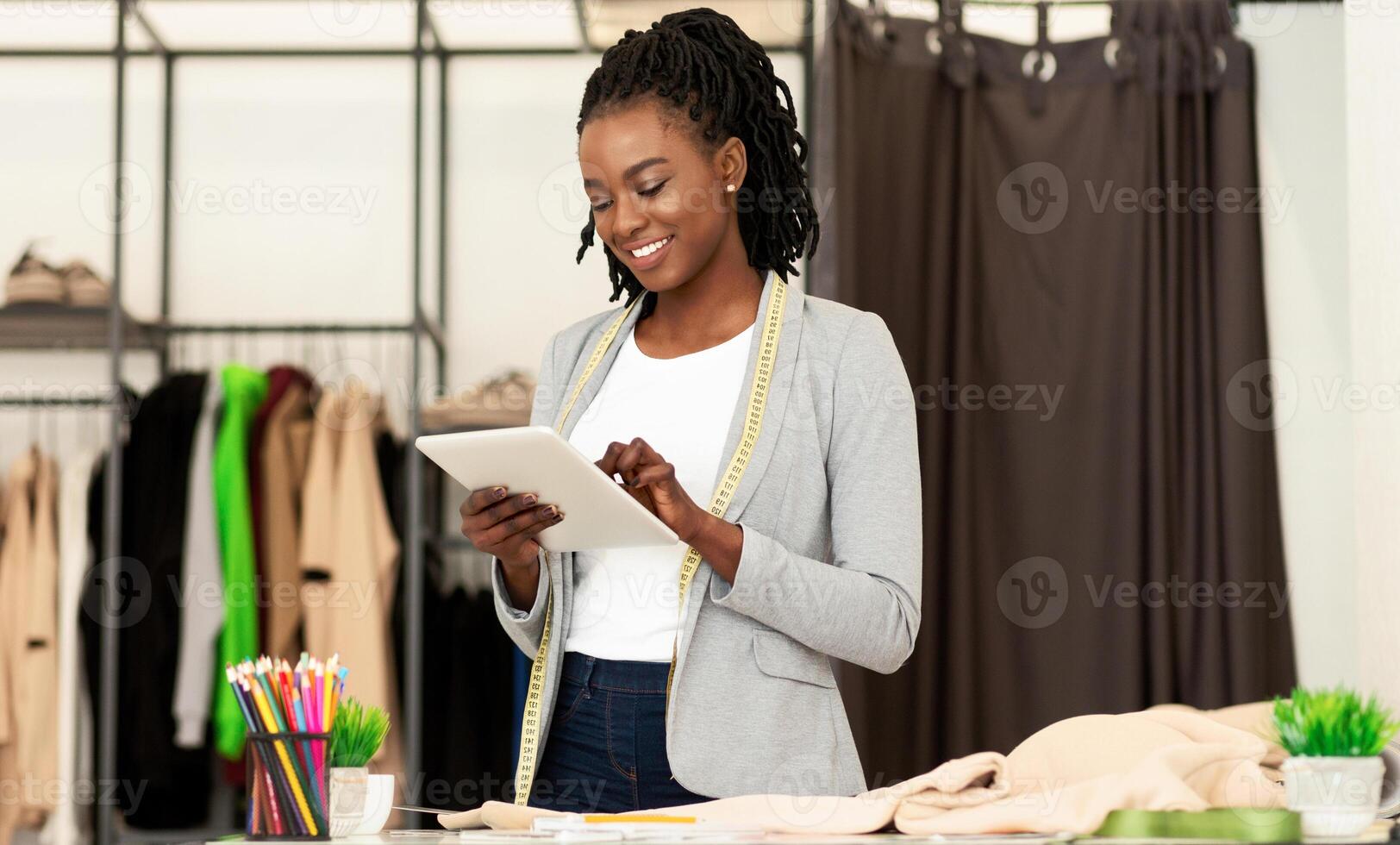 Cheerful Afro Fashion Designer Using Tablet Computer Browsing Internet In Dressmaking Studio. Empty Space photo