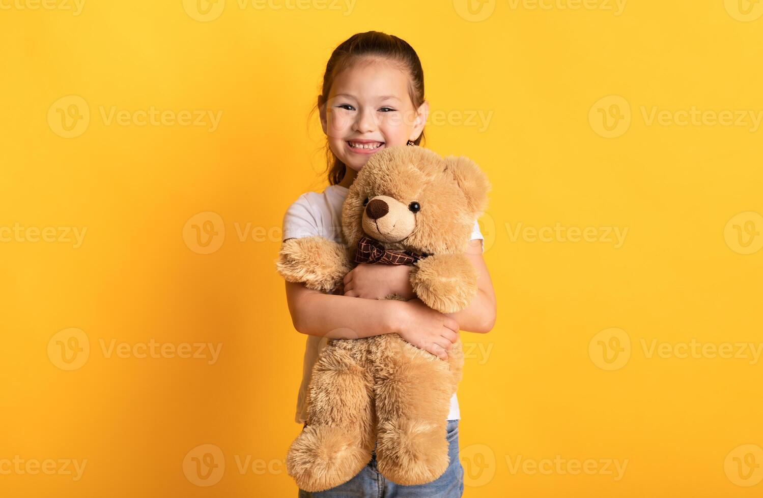 Portrait of adorable little asian girl holding teddy bear in hands standing isolated over yellow studio wall, copy space photo