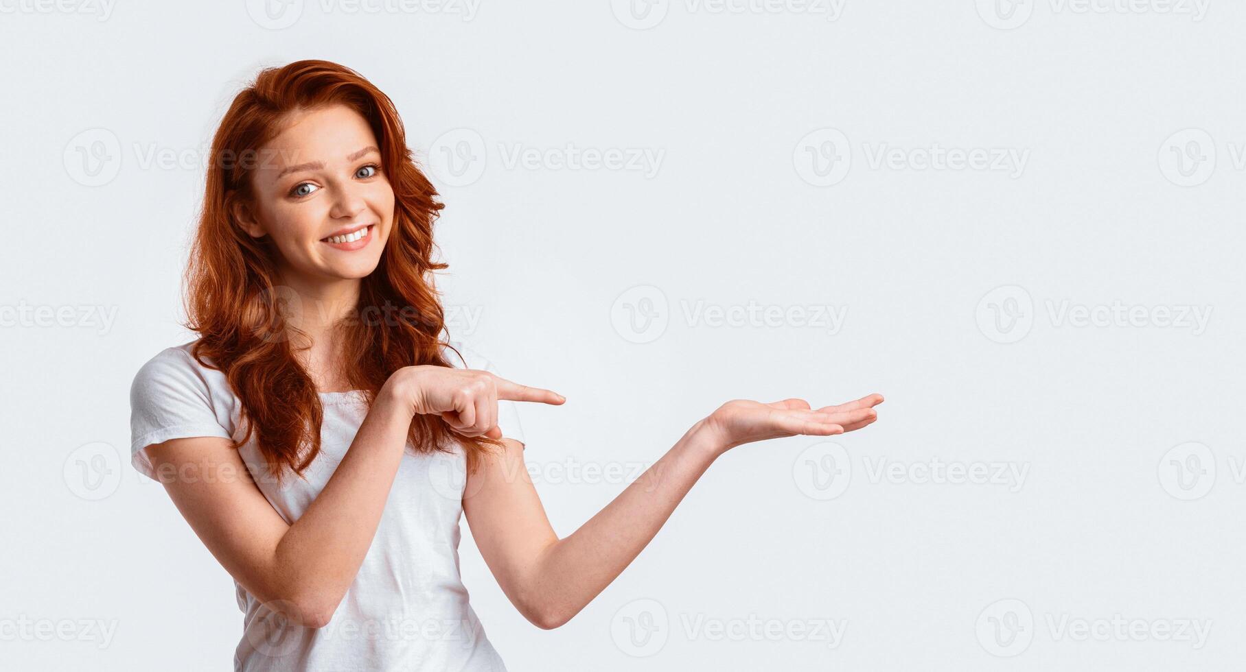Look At This. Ginger-Haired Girl Holding Something And Pointing At Invisible Object On Hand Standing Over White Studio Background. Panorama photo