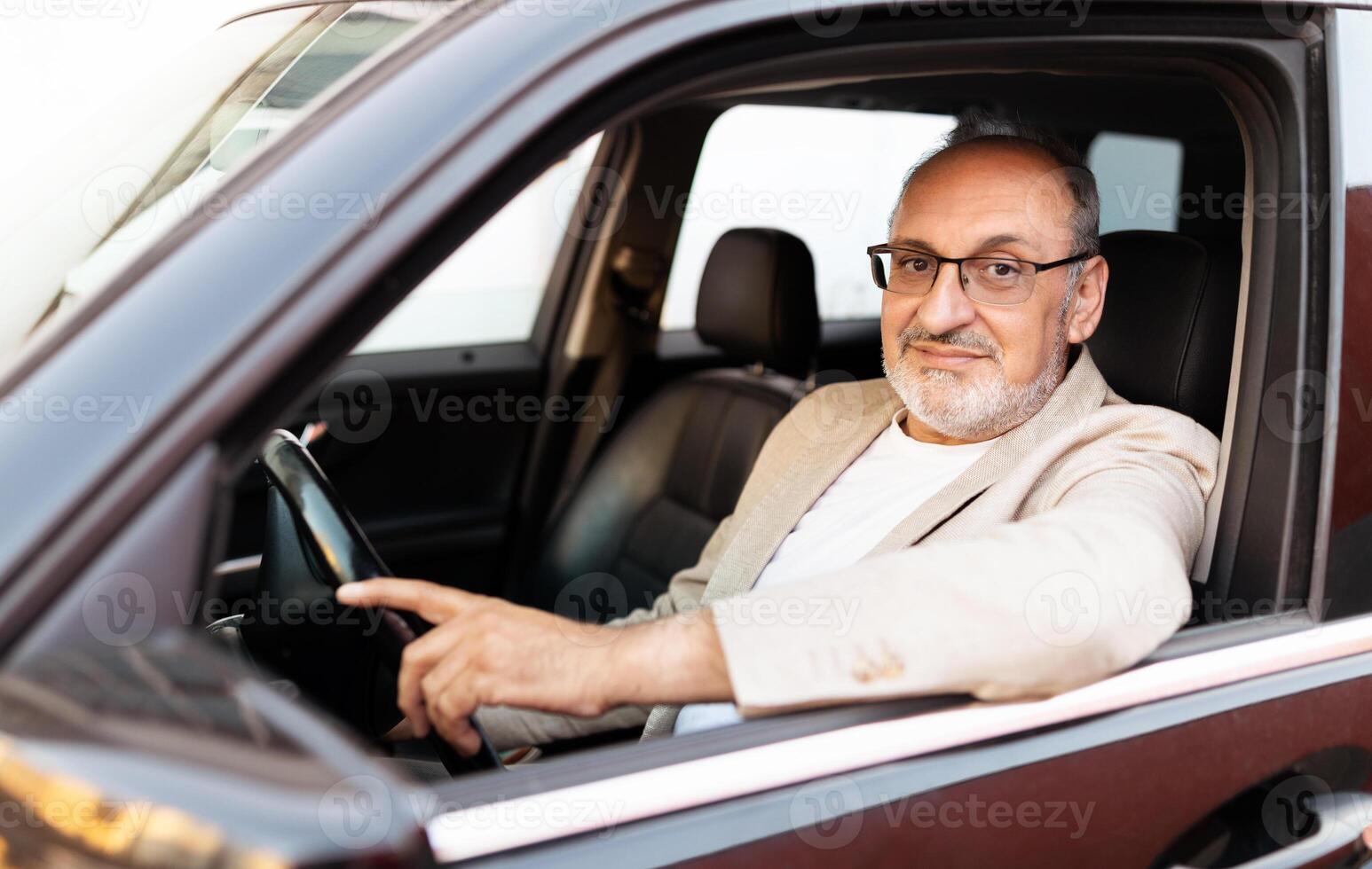 A confident man with glasses is seated in the driver's seat of a car, pointing outside as he looks at the camera. The warm daylight highlights his relaxed demeanor. photo