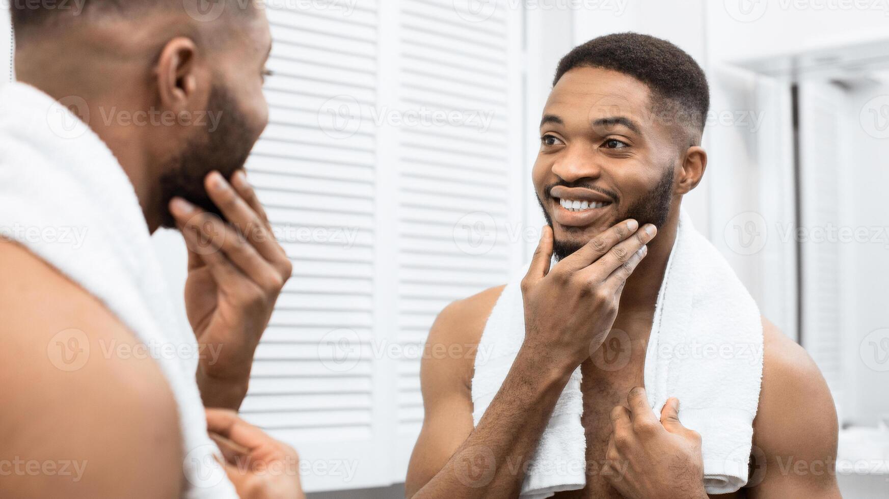 Happy afro man with bare torso touching beard looking in mirror at bathroom. Modern man in bathroom concept photo
