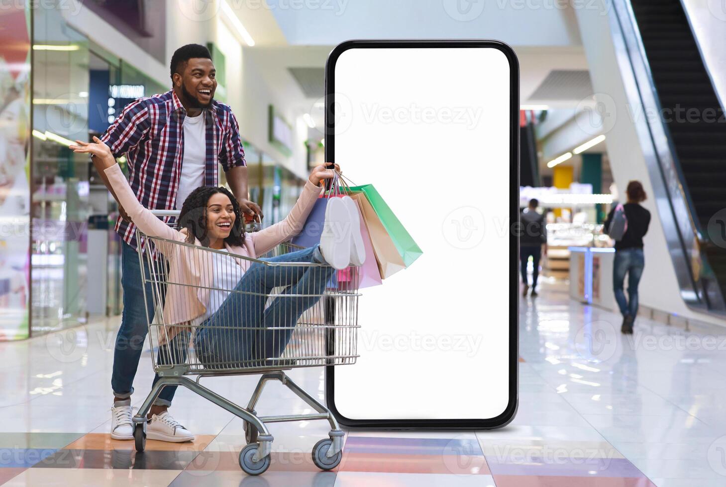 A joyful black couple engages in a fun shopping experience at a vibrant mall. The man pushes a shopping cart with the woman sitting inside, surrounded by colorful bags and a large blank screen nearby. photo