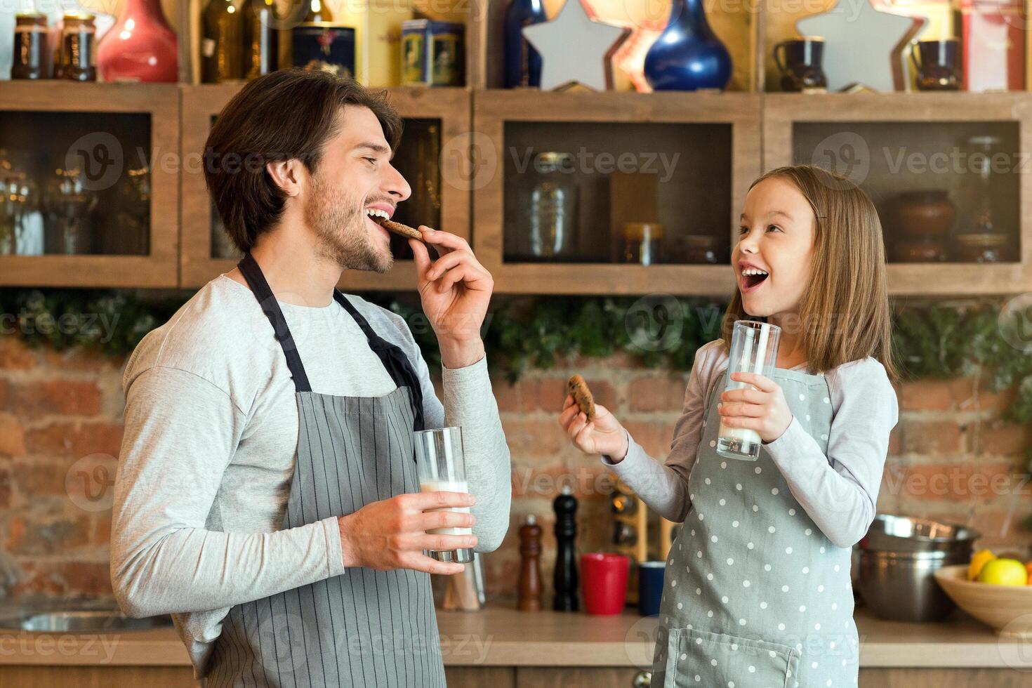Snack Time. Cheerful dad and little daughter eating fresh baked cookies and drinking milk in kitchen, enjoying homemade pastry, wearing aprons and smiling to each other, having fun at home, copy space photo