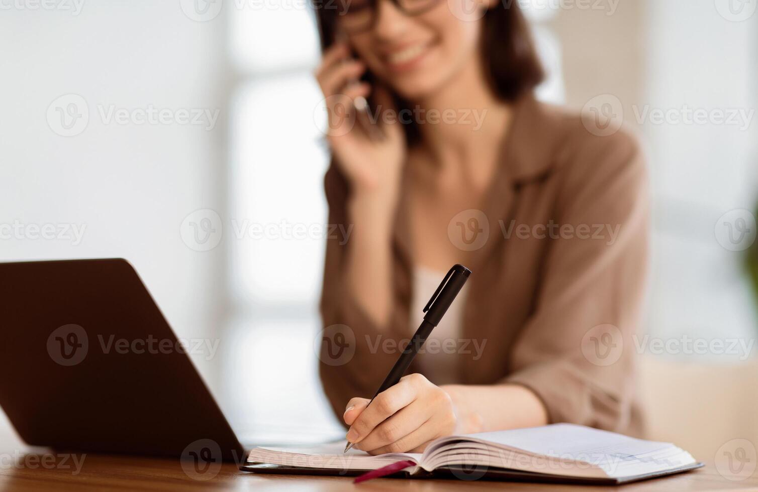 Taking Notes. Young secretary sitting at desk with laptop computer and talking on mobile phone, writing in notebook photo