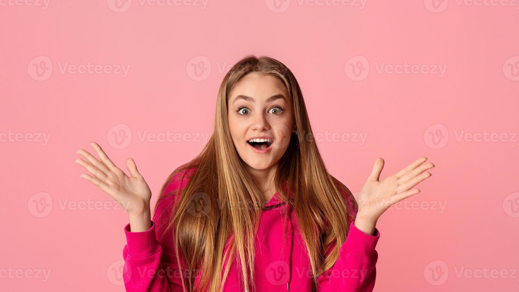 Expression of surprise and happiness. Emotional teen girl with open mouth spreads hands isolated on pink background photo